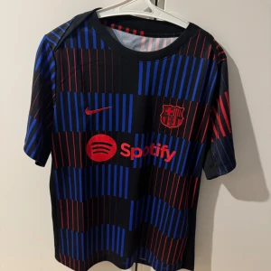 FC Barcelona träningströja Nike - Säljer en FC Barcelona träningströja från Nike med Spotify-logga. Tröjan är svart med vertikala ränder i blått och rött, klubbmärke och Nike-logga på bröstet. Kortärmad och tillverkad i lätt funktionsmaterial.