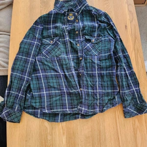 Grön och blå rutig skjorta  - Säljer en klassisk grön och blå rutig skjorta med knappar framtill och två bröstfickor. Skjortan har lång ärm och är perfekt för en avslappnad stil. Märkeslogga på insidan av kragen.
