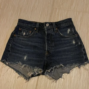 Mörkblå jeansshorts från Levi's 501 - Säljer ett par mörkblå jeansshorts från Levi's 501 med fransig kant och slitna detaljer. Klassisk femficksmodell med låg midja. Perfekta för en avslappnad stil. De är i bra skick, är nya och har inte används. Storlek W24