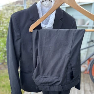 Svart kostym från H&M strl 152 - Säljer en klassisk svart kostym från H&M med kavaj och matchande byxor. Kavajen har smal passform, enkla slag och två fickor framtill. Byxorna har raka ben och bälteshällor. Perfekt för formella tillfällen. Använd 1 gång. Storlek 152.