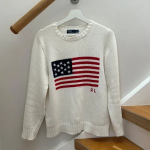Vit stickad tröja med amerikansk flagga från Polo Ralph Lauren - Vit stickad tröja från Polo Ralph Lauren med amerikansk flagga och RL-broderi framtill. Rund halsringning och ribbade muddar vid ärmslut och nederkant. Perfekt för en klassisk och avslappnad stil. Helt ny i princip använt den en gång.