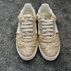 Valentino sneakers med guldglitter - Säljer ett par snygga Valentino sneakers täckta i guldglitter med vita detaljer och vita snören. Skorna har en chunky vit sula och små nitar baktill. Perfekta för dig som vill sticka ut med lite extra bling!