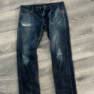 Mörkblå jeansbyxor med slitningar - Säljer ett par mörkblå jeansbyxor med slitningar på båda benen och klassisk femficksdesign. Jeansen har en rak passform och tydliga tvättade detaljer för en cool, använd look. Perfekta för dig som gillar en avslappnad stil.