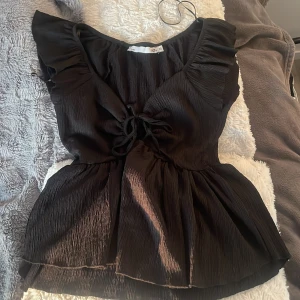 Svart blus med volangärmar och knytning - Säljer en svart blus med volangärmar och knytning framtill. Blusen har en lätt rynkad struktur och v-ringning. Perfekt för dig som vill ha en feminin och trendig topp.
