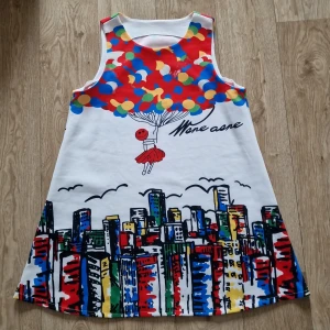 Färgglad ärmlös klänning med stadsmotiv - Säljer en vit ärmlös klänning med färgglatt tryck av ballonger och stadssilhuett. Klänningen har en lekfull design med en tecknad figur som håller i ballonger. Perfekt för dig som gillar unika och färgstarka plagg!