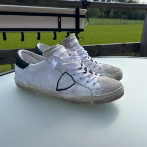 Vita sneakers från Philippe Model - Säljer ett par vita Philippe Model sneakers med svarta detaljer på hälen och grå mocka framtill. Skorna har snörning och en platt sula. Klassisk design med märkets logga på sidan och bak på hälen.