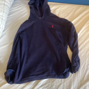 Mörkblå hoodie från Polo Ralph Lauren - Säljer en mörkblå hoodie från Polo Ralph Lauren med den klassiska röda broderade loggan på bröstet. Tröjan har lång ärm och huva, perfekt för en avslappnad stil. Materialet är mjukt och skönt bomullstyg.