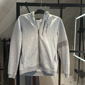 Grå hoodie från Sail Racing - Säljer en grå hoodie från Sail Racing med dragkedja framtill och på bröstfickan. Tröjan har huva och logga på bröstet samt på huvan. Perfekt för en sportig och avslappnad stil. Säljer då den är för liten. Säljer såpass billigt för att den har en defekt på höger armen. Så min mormor har sytt igen hålet. Hör av er vid frågor! 