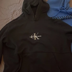 Svart hoodie från Calvin Klein Jeans - Svart hoodie från Calvin Klein Jeans med vit logga på bröstet. Klassisk huva med dragsko och känguruficka framtill. Perfekt för en avslappnad och stilren look.