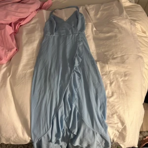 Ljusblå långklänning med volang från SHEIN - Säljer en ljusblå långklänning från SHEIN med smala axelband, omlott-design och volangdetaljer framtill. Klänningen har v-ringad hals och snörning i ryggen för en snygg passform. Perfekt för sommarens tillfällen!