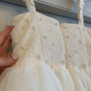 Vit pärlprydd klänning - Säljer en superfin vit klänning med pärlprytt liv och justerbara axelband. Överdelen är täckt av små pärlor och har en markerad byst, medan kjolen är i flera lager organza som ger en fluffig känsla. Perfekt för speciella tillfällen!