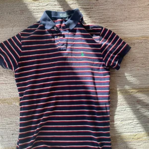  pikétröja från Polo Ralph Lauren - Mörkblå pikétröja med röda och vita ränder från Polo Ralph Lauren. Inga defekter mycket bra skick. 