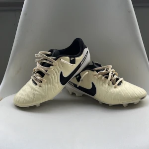 Vita Tienpos fotbollsskor med svarta detaljer - Säljer ett par vita Nike fotbollsskor med svarta swoosh-loggor och snörning. Skorna har rund tå och platt sula med dobbar för bra grepp på planen. Insidan är svart och materialet känns syntetiskt. Skorna har använts ett fåtal träningar. 