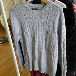 Grå kabelstickad tröja från East West - Säljer en klassisk grå kabelstickad tröja från East West Athletic Supply Co. Tröjan har rund halsringning och långa ärmar. Perfekt för lager-på-lager och enkel att matcha med olika outfits. Stl S. Fint skick! 