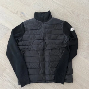 Moncler cardigan  - Svart Moncler-jacka med quiltad front och rygg i syntetmaterial samt stickade ärmar. Jackan har dragkedja framtill och två fickor med dragkedja. Klassisk Moncler-logga på vänster ärm. Perfekt för lager-på-lager.