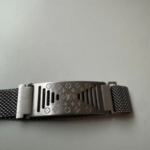 Lv Armabnd  - Stilrent armband i silverfärgad metall med meshband och en platta dekorerad med Louis Vuitton-logga och ikoniska mönster. Perfekt accessoar för att lyfta din outfit med en lyxig touch. Ett väldigt sällsynt armband som passar alla handleder med hjälp av magnet spännet.