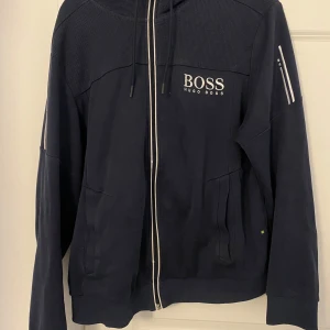 Mörkblå hoodiejacka från Hugo Boss - Snygg mörkblå hoodiejacka från Hugo Boss med vit logga på bröstet och dragkedja framtill. Jackan har huva med dragsko, ribbade muddar och en ficka med dragkedja på ärmen. Perfekt för en stilren och sportig look.