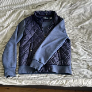 Blå quiltad Cardigan från Polo Ralph Lauren - Snygg blå quiltad cardigan från Polo Ralph Lauren med dragkedja och tryckknappar. Jackan har ribbade muddar och en stilren design som passar perfekt för både höst och vår. Den är tillverkad i ett syntetmaterial som ger en skön känsla. 🤩