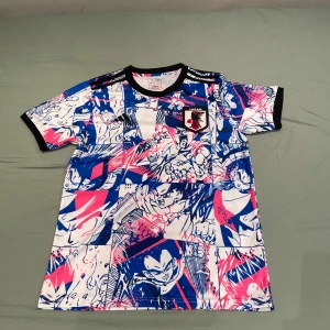 Japan fotbollströja anime-mönster - Säljer en Japan fotbollströja från Adidas med unikt anime-inspirerat mönster i blått, vitt och rosa. Tröjan har korta ärmar, svart krage och detaljer samt Japan-emblem på bröstet. Perfekt för dig som gillar både fotboll och anime!