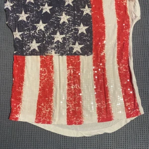 T-shirt med USA-flagga och paljetter - Cool t-shirt med amerikanska flaggan i sliten look framtill och paljettdetaljer som ger extra bling. Toppen är vit med röda och blå inslag, har rund hals och är kortärmad. Perfekt för dig som vill sticka ut lite extra.