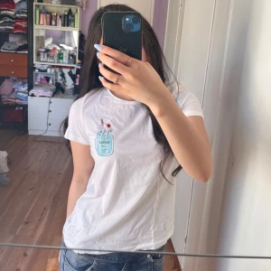 Vit t-shirt med tryck från Vero Moda - Vit t-shirt från Vero Moda med ett litet tryck av en blå dryckesflaska på bröstet. Klassisk rund halsringning och korta ärmar. Perfekt till jeans för en avslappnad look.