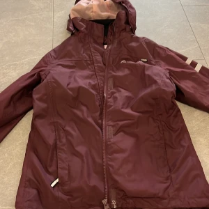 Vinröd regnjacka från On The Peak - Säljer en vinröd regnjacka från On The Peak med huva och dragkedja framtill. Jackan har reflekterande detaljer på ärmen och är tillverkad i vattentätt material (Aquatex). Perfekt för blöta dagar.