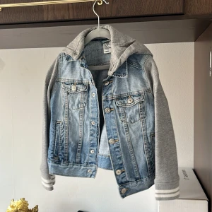 Jeansjacka med grå ärmar och huva från H&M - Snygg jeansjacka från H&M med ljusblå denim och grå sweatshirt-ärmar samt huva. Jackan har knappar framtill, två bröstfickor och randiga muddar vid ärmsluten. Perfekt för en avslappnad streetstil.