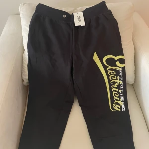 Svarta mjukisbyxor från WARP med gul text - Svarta mjukisbyxor från WARP med stor gul text 'Electricity' och vit text på ena benet. Byxorna har resår i midjan och mudd vid bensluten. Perfekta för en avslappnad stil och riktigt sköna att ha på sig.