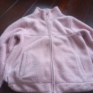 Ljusrosa fleecejacka med dragkedja - Mysig ljusrosa fleecejacka med hög krage och hel dragkedja framtill. Jackan har två fickor på sidorna och är perfekt för kyliga dagar. Mjuk och fluffig känsla som håller dig varm.