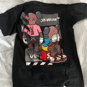 Svart Off-White t-shirt med KAWS-motiv - Svart t-shirt från Off-White med stor färgglad KAWS-inspirerad print på ryggen och en sneaker-grafik på bröstet. Klassisk passform och rund halsringning. Tillverkad i 100% bomull-köpt i på plats i en offwhite butik. Den är i storlek S men passar även M.