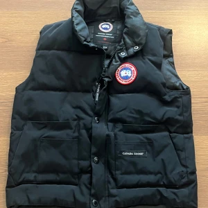 Svart dunväst från Canada Goose - Säljer en svart dunväst från Canada Goose med klassisk logotyp på bröstet och två stora fickor framtill. Västen har hög krage, knappar och dragkedja. Perfekt för kyliga dagar. Lite liten i storlek