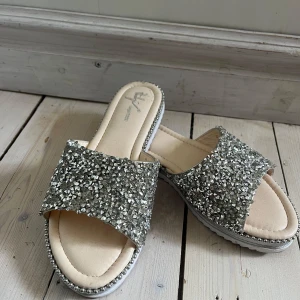 Glittriga sandaler i silver  - Säljer ett par sandaler med bred rem täckt av glittriga silverpaljetter. Sköna sulor, endast använda en gång. Storlek 38