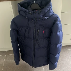 Mörkblå dunjacka från Polo Ralph Lauren - Säljer en mörkblå dunjacka från Polo Ralph Lauren med huva och dragkedja framtill. Jackan har en bröstficka med dragkedja och den klassiska röda loggan broderad på bröstet. Perfekt för kyliga dagar.