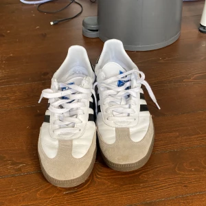 Adidas Samba sneakers i vitt och grått - Klassiska Adidas Samba sneakers med vit ovandel, grå mockadetaljer och svarta ränder på sidorna. Skorna har snörning, gummisula och den ikoniska Samba-loggan i guld på sidan. Perfekta för dig som gillar retrostil och streetwear.