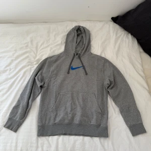 Grå Nike hoodie  - Säljer en grå hoodie från Nike med blå swoosh-logga på bröstet. Tröjan har huva med dragsko och en stor magficka framtill. Perfekt för en avslappnad stil och enkel att matcha med det mesta.