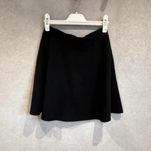 Svart kortkjol - Enkel och stilren svart kortkjol med lätt utställd passform. Perfekt att matcha med olika toppar för en clean look. Kjolen har en mjuk och följsam känsla.