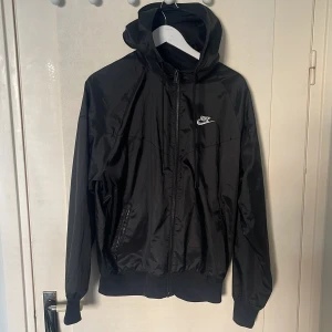 Svart vindjacka från Nike med huva - Säljer en svart vindjacka från Nike med huva och dragkedja framtill. Jackan har två fickor med dragkedja och ribbade muddar vid ärmslut och nederkant. Klassisk Nike-logga på bröstet och lätt material.