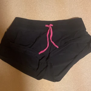 Svarta shorts från Rebecca Bonbon med rosa snörning - Svarta shorts från Rebecca Bonbon med rosa snörning i midjan och tryckt logga bak. Mjukt material och normal passform, perfekta för träning eller chill. Snygg kontrast mellan svart tyg och rosa detaljer.