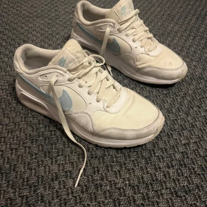 Nike Air Max  - Säljer ett par vita Nike Air sneakers med ljusblå swoosh och detaljer. Skorna har snörning, rund tå och platt sula. Perfekta för dig som gillar klassisk och sportig stil. Passar till många olika outfits och är riktigt bekväma.