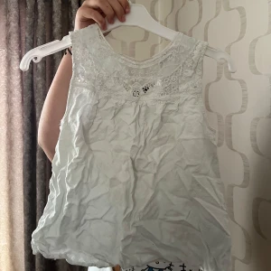 Vit ärmlös blus med spetsdetaljer - Söt vit blus med spets upptill och ärmlös design. Blusen har rund halsringning och fina broderade detaljer vid axlar och bröst. Perfekt för varma dagar och enkel att matcha med olika underdelar.