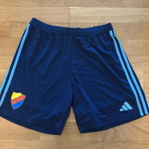 Blå fotbollsshorts från Adidas x Djurgården - Mörkblå fotbollsshorts med ljusblå ränder längs sidorna och Djurgårdens IF klubbmärke på ena benet. Tillverkade i lätt syntetmaterial med elastisk midja och Aeroready-teknologi för bra ventilation. Perfekta för träning eller match.