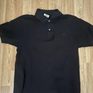 Svart pikétröja från Lacoste - Klassisk svart pikétröja från Lacoste med krage och två knappar framtill. Tröjan har korta ärmar och är tillverkad i ett mjukt bomullsmaterial. Perfekt för en stilren och avslappnad look.
