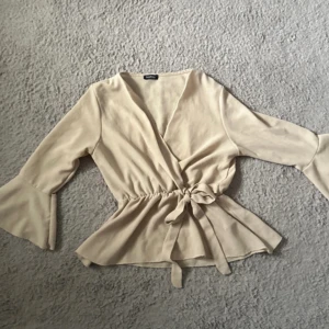 Beige omlottblus med volangärm - Säljer en beige omlottblus med knyt i midjan och volangdetaljer vid ärmslut och nederkant. Blusen har v-ringning och långa ärmar, perfekt för en trendig och stilren look. Använd ett fåtal gånger, inga defekter 