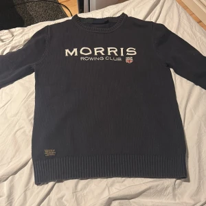 Mörkblå stickad tröja från Morris - Säljer en mörkblå stickad tröja från Morris med texten 'Morris Rowing Club' broderad på bröstet. Tröjan har rund halsringning och långa ärmar med ribbade muddar och nederkant. Perfekt för en stilren och sportig look.