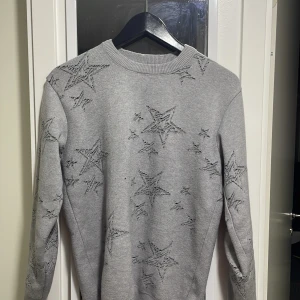 Grå stickad tröja med stjärnmönster från Kenzo - Säljer en grå stickad tröja från Kenzo med broderade stjärnor över hela tröjan. Rund halsringning och långa ärmar. Perfekt för dig som vill ha något unikt och snyggt i garderoben.