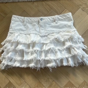 Vit volangkjol från Monki - Supersöt vit kjol från Monki med flera lager volanger och fransiga kanter. Kjolen har klassisk jeansmidja med knapp och hällor för skärp. Perfekt för dig som vill sticka ut med en lekfull och trendig look.