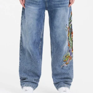 Edhardy jeans med drakbroderi - Säljer ett par baggy jeans med stort färgglatt drakbroderi längs ena benet och broderade detaljer baktill. Jeansen har en lös passform och klassisk blå tvätt. Perfekta för dig som vill sticka ut med coola detaljer och streetstyle.