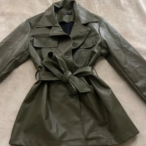 Olivgrön skinnjacka med bälte från SHEIN - Snygg olivgrön skinnjacka från SHEIN med markerad krage och bälte i midjan. Jackan har långa ärmar och en klassisk design som ger en stilren look. Perfekt för dig som vill ha en trendig jacka med lite edge.