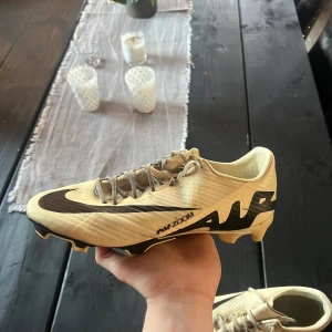 Nike Air Zoom Mercurial fotbollsskor beige och svart - Säljer ett par Nike Air Zoom Mercurial fotbollsskor i beige med svarta detaljer och stor Nike-logga på sidan. Skorna har snörning och är designade för spel på gräs. Perfekta för dig som vill sticka ut på planen.