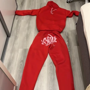 Syna world tracksuit red  - Säljer en röd hoodie med vit tryckdetalj på huvan. Hoodien har långa ärmar, magficka och snörning i halsen. Perfekt för en avslappnad stil.